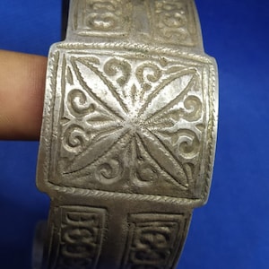 May include: A silver bracelet with intricate, embossed designs. The bracelet is made of a single piece of metal and has a square-shaped design in the center.