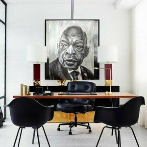 以下が含まれることがあります： スーツを着た男性の白黒の肖像画が、黒い革張りのオフィスチェアのある木製デスクの上に飾られています。デスクの両側には、金色の脚が付いた黒い椅子が2つあります。部屋は白い壁と白い天井です。