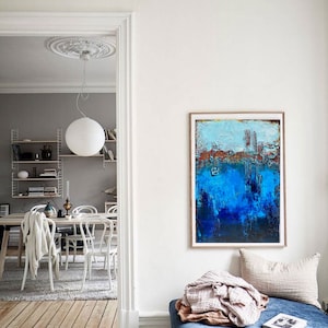 Könnte beinhalten: Ein blaues und weißes abstraktes Gemälde in einem Holzrahmen hängt an einer weißen Wand über einer blauen Samtbank mit einer Wolldecke und einem Buch.