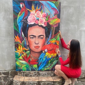 Pu&ograve; includere: Un dipinto colorato di Frida Kahlo con pappagalli e fiori. Il dipinto &egrave; su tela ed &egrave; tenuto da una persona.