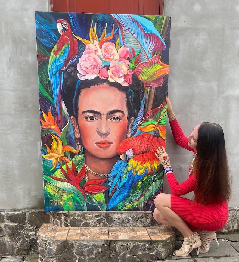 Pu&ograve; includere: Un dipinto colorato di Frida Kahlo con pappagalli e fiori. Il dipinto &egrave; su tela ed &egrave; tenuto da una persona.