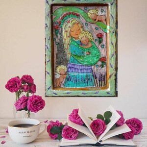 May include: A framed painting of a woman holding a baby with two angels above them. The painting is on a wooden background with a green, blue, and purple color scheme. The painting is surrounded by a white frame with a lavender border.