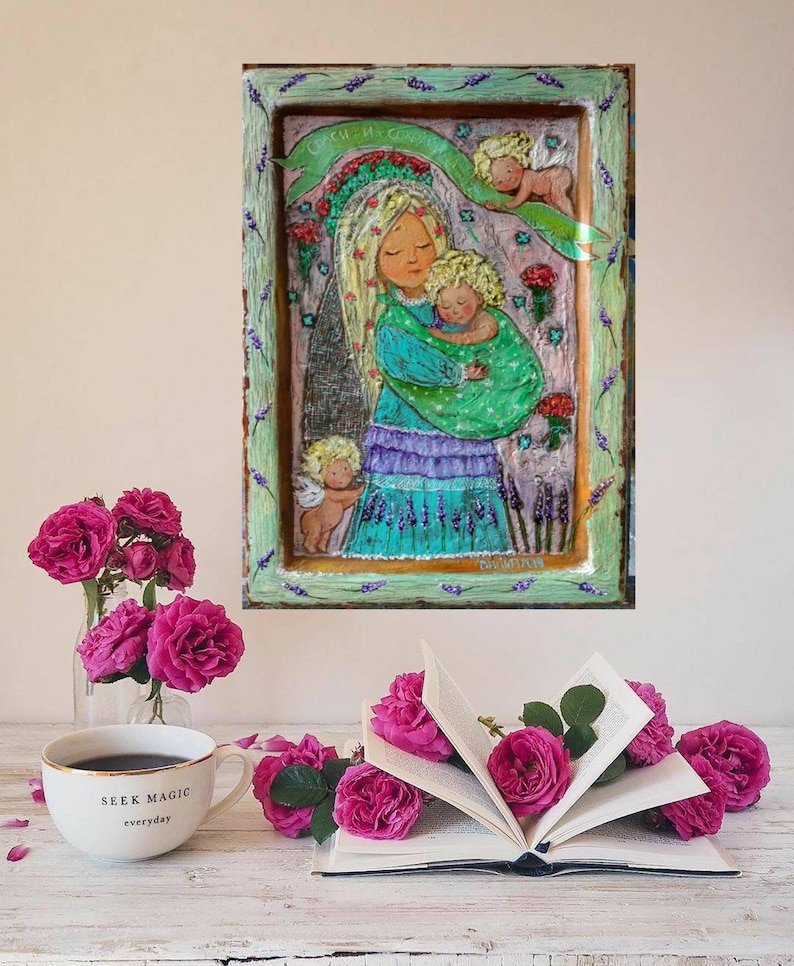 May include: A framed painting of a woman holding a baby with two angels above them. The painting is on a wooden background with a green, blue, and purple color scheme. The painting is surrounded by a white frame with a lavender border.