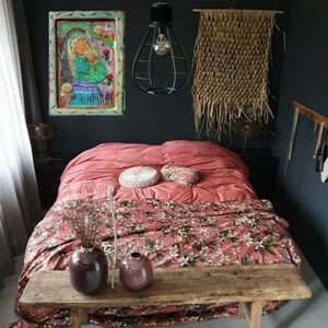 May include: A bedroom with a pink floral duvet cover, a wooden bench, and two purple vases. A framed painting of a woman and child hangs on the wall above the bed. A woven wall hanging is on the wall to the right of the bed.