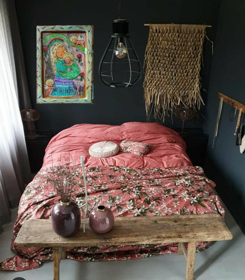 May include: A bedroom with a pink floral duvet cover, a wooden bench, and two purple vases. A framed painting of a woman and child hangs on the wall above the bed. A woven wall hanging is on the wall to the right of the bed.