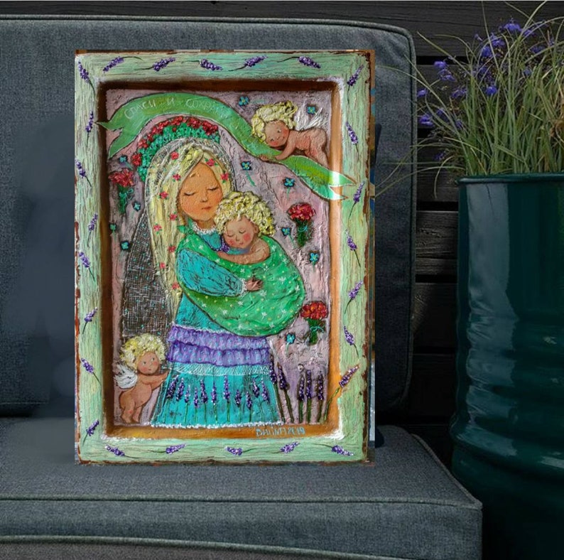 May include: A framed painting of a woman holding a child. The woman is wearing a blue and purple dress and a crown of flowers. The child is wearing a green dress. The painting is surrounded by a light green frame with lavender flowers.