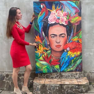 Pu&ograve; includere: Un dipinto colorato di Frida Kahlo con una corona di fiori e pappagalli. Il dipinto &egrave; su tela ed &egrave; tenuto da una persona.