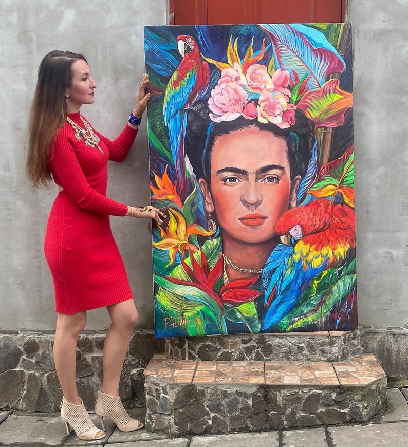 Pu&ograve; includere: Un dipinto colorato di Frida Kahlo con una corona di fiori e pappagalli. Il dipinto &egrave; su tela ed &egrave; tenuto da una persona.