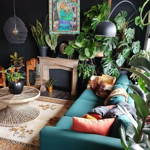 May include: A teal green sofa with a variety of throw pillows in a living room with a fireplace and many plants. The artwork above the fireplace is a painting of a woman holding a child.