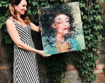 May include: A woman in a black and white striped dress holds a painting of a woman's face. The painting is done in a mosaic style with shades of blue, green, and brown.