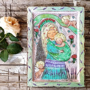 May include: A framed painting of a woman holding a child. The woman is wearing a blue and green dress and a crown of flowers. The child is wearing a green and white dress. The painting is surrounded by a green frame with lavender flowers.