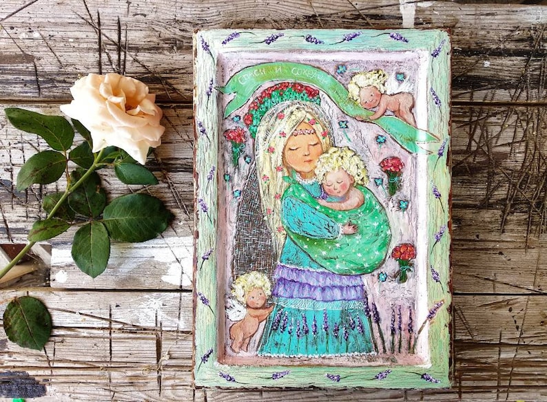 May include: A framed painting of a woman holding a child. The woman is wearing a blue and green dress and a crown of flowers. The child is wearing a green and white dress. The painting is surrounded by a green frame with lavender flowers.