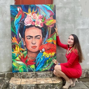 Pu&ograve; includere: Un dipinto colorato di Frida Kahlo circondata da fiori tropicali e pappagalli. Il dipinto &egrave; su tela ed &egrave; tenuto da una persona con un vestito rosso.