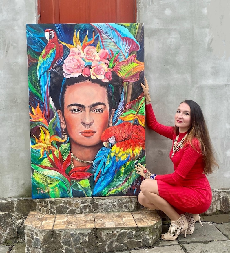 Pu&ograve; includere: Un dipinto colorato di Frida Kahlo circondata da fiori tropicali e pappagalli. Il dipinto &egrave; su tela ed &egrave; tenuto da una persona con un vestito rosso.