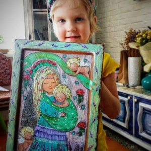 May include: A framed painting of a woman holding a baby with two angels on either side. The painting is in a green frame with a lavender border. The painting is titled "Спаси и Сохрани" which translates to "Save and Preserve".