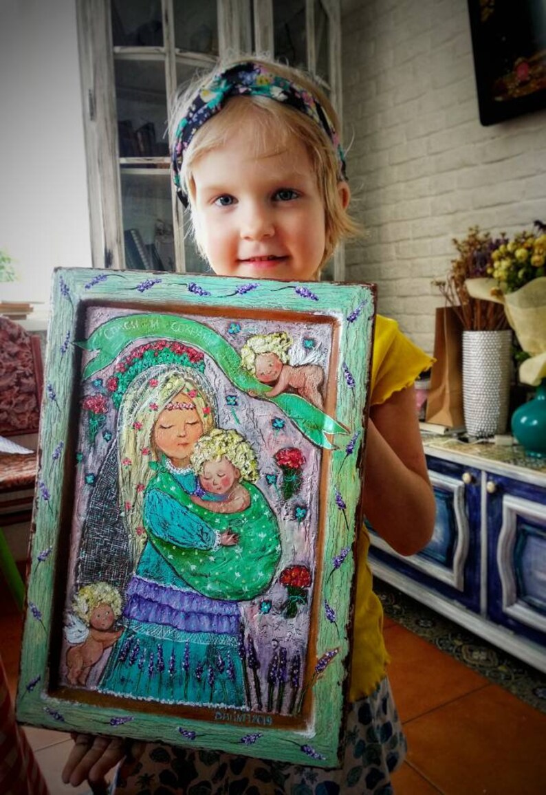 May include: A framed painting of a woman holding a baby with two angels on either side. The painting is in a green frame with a lavender border. The painting is titled "Спаси и Сохрани" which translates to "Save and Preserve".