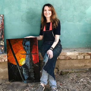 May include: A woman in a black shirt and denim overalls is holding a painting of a man with a beard and a red and orange background. The painting is framed and has a black border.