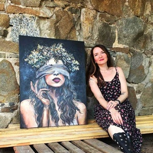 May include: A painting of a woman with a blindfold on, wearing a crown of flowers. The woman is looking to the side with a thoughtful expression. The painting is on a wooden surface, and a woman is sitting on the surface behind the painting.