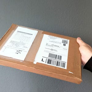 May include: A rectangular brown cardboard shipping box with two white adhesive labels attached to the top surface. The labels feature printed text, barcodes, and a QR code. The box is held at the side, showing a flat, thin profile and tape.
