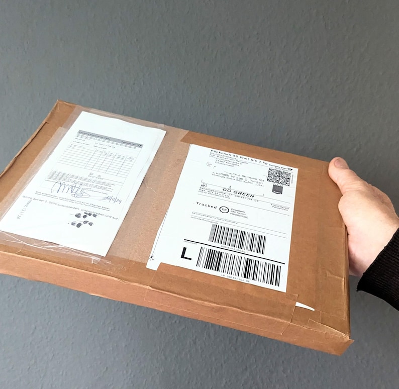 May include: A rectangular brown cardboard shipping box with two white adhesive labels attached to the top surface. The labels feature printed text, barcodes, and a QR code. The box is held at the side, showing a flat, thin profile and tape.