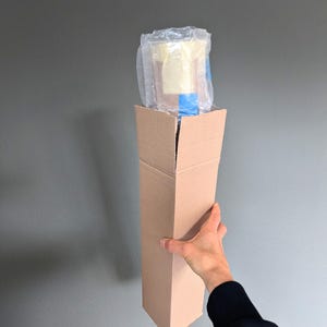 May include: A tall, rectangular cardboard box, held by a person's hand, contains a wrapped item. The item is encased in clear plastic and blue tape. The box is plain brown and open at the top.