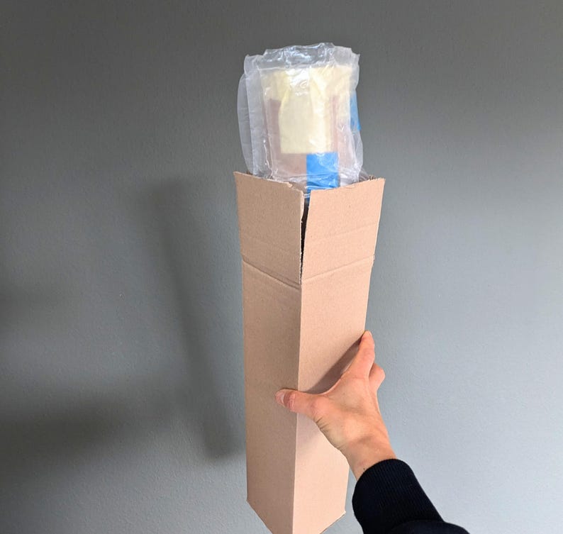 May include: A tall, rectangular cardboard box, held by a person's hand, contains a wrapped item. The item is encased in clear plastic and blue tape. The box is plain brown and open at the top.