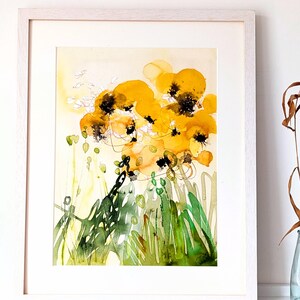 May include: Abstract watercolor painting of yellow flowers with black centers and green foliage. The artwork is framed in a white wooden frame. The painting has a light yellow background with splashes of color.
