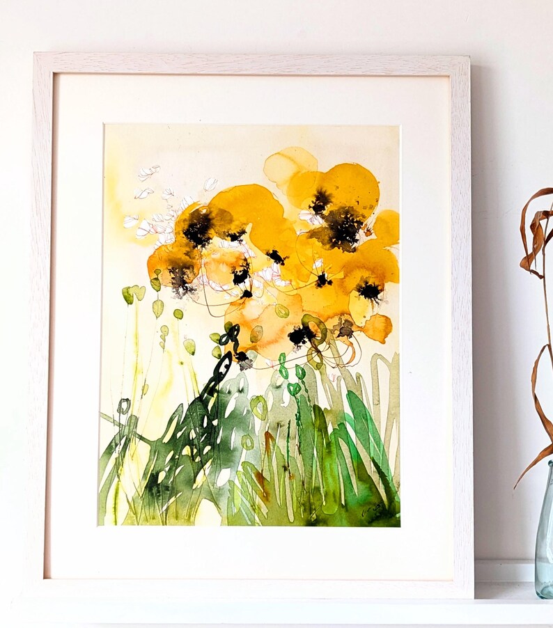 May include: Abstract watercolor painting of yellow flowers with black centers and green foliage. The artwork is framed in a white wooden frame. The painting has a light yellow background with splashes of color.