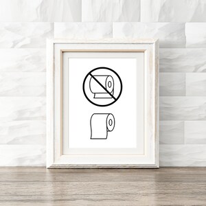 May include: A white framed print with a black and white illustration of a toilet paper roll with a red circle and diagonal line through it. Below the circle is a black and white illustration of a toilet paper roll.