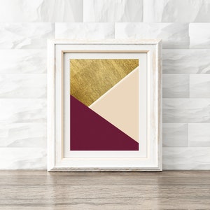 Könnte beinhalten: Ein gerahmter Kunstdruck mit abstraktem Design in einem weißen Holzrahmen. Das Kunstwerk zeigt ein Farbblockdesign in Gold, Creme und Burgunderrot. Der Druck wird auf einer Holzoberfläche vor einer weißen Backsteinmauer präsentiert.