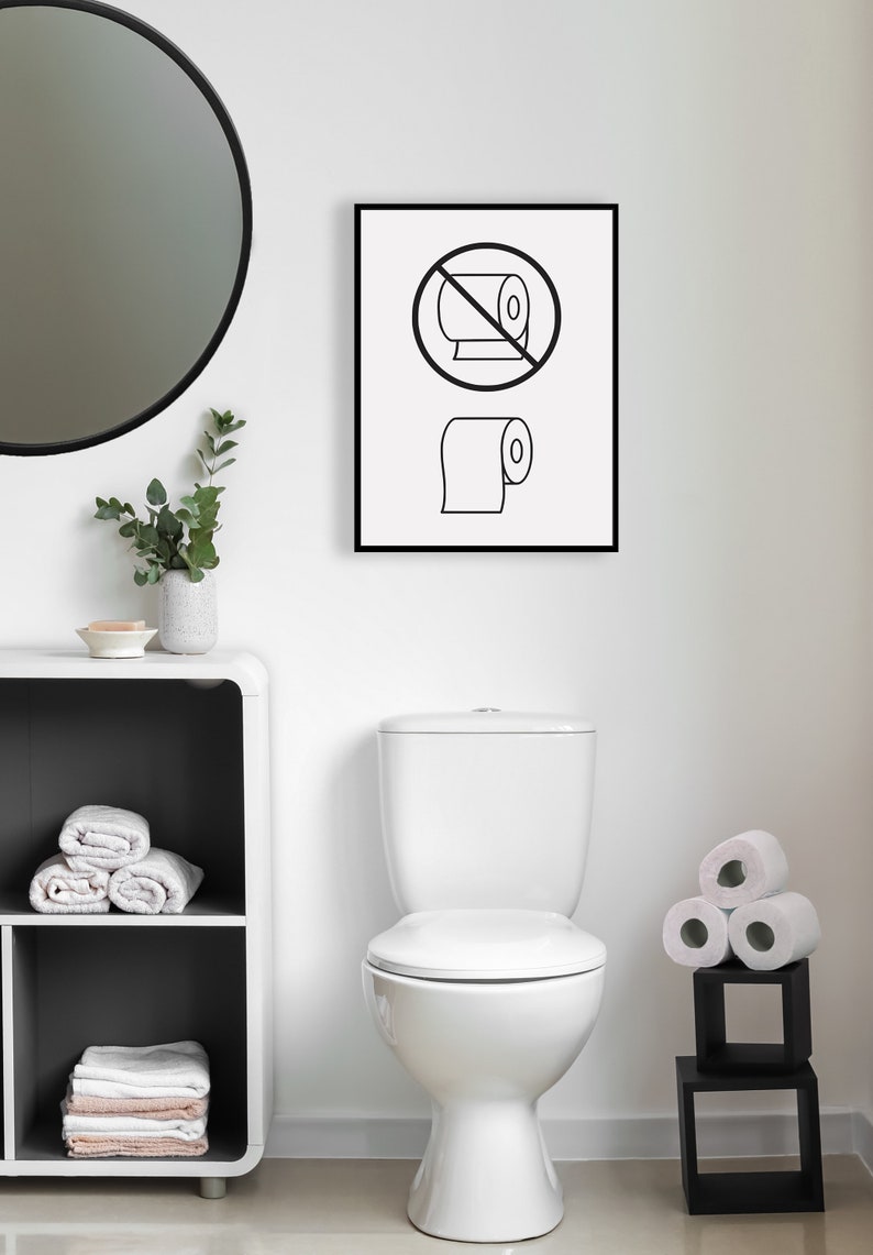 May include: A white toilet with a closed lid in a bathroom with a black framed sign that says "Do not roll toilet paper" with a black circle and a line through a roll of toilet paper and a roll of toilet paper below it. There is a white round mirror on the wall, a white shelf with towels, and a black shelf with rolls of toilet paper.