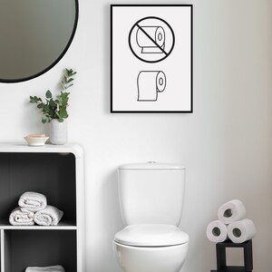 May include: A white toilet with a closed lid in a bathroom with a black framed sign that says "Do not roll toilet paper" with a black circle and a line through a roll of toilet paper and a roll of toilet paper below it. There is a white round mirror on the wall, a white shelf with towels, and a black shelf with rolls of toilet paper.