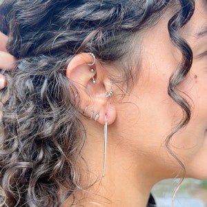 May include: Close-up of an ear with multiple silver earrings. The ear has several piercings, each holding a different style of earring, including hoops, studs, and a long, dangling bar earring. Curly hair frames the ear.