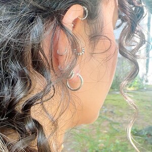 Peut inclure: Gros plan sur une oreille ornée de plusieurs boucles d'oreilles argentées. Les boucles d'oreilles comprennent de petits cerceaux, un design perlé et un cerceau plus grand. Des cheveux bouclés foncés encadrent l'oreille, avec un arrière-plan vert flou.