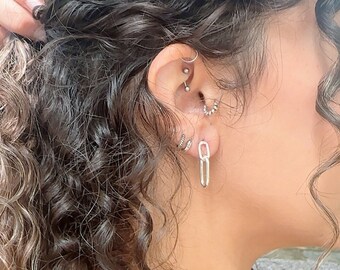 May include: Close-up of an ear with multiple piercings, including a silver chain link earring. The ear is surrounded by dark, curly hair. The skin tone is light brown.