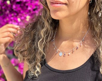 May include: Silver necklace with dangling charms and small beads in shades of purple, white, and gray. The necklace has a small, round pendant. The model is wearing a black tank top.
