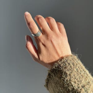 Peut inclure: Une bague argentée au design incurvé est portée au doigt. La bague est lisse et réfléchissante, captant la lumière. L'arrière-plan est gris clair, avec un aperçu d'une manche de pull vert olive texturée.