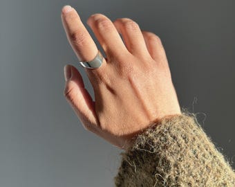 May include: A silver ring with a curved design is worn on a finger. The ring is smooth and reflective, catching the light. The background is a soft gray, with a glimpse of a textured, olive-green sweater sleeve.