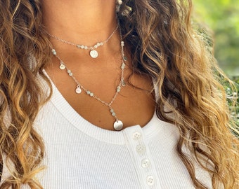 May include: A layered silver necklace with small, round, light green beads and dangling silver disc charms. The necklace is worn over a white top. The background is blurred, with green foliage visible.