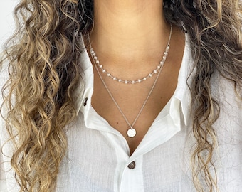 May include: A layered necklace with a delicate design. The top layer features small, white beads, while the lower layer has a circular pendant. The necklaces are silver-toned and worn over a white shirt.