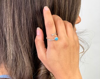 May include: A silver ring with a small, round, light blue stone and a smaller clear stone. The ring is on a finger, with the hand and arm visible. The ring is a piece of jewelry.