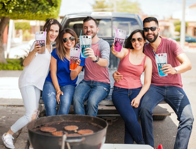 May include: A group of friends enjoying drinks from colourful pouches with the word "TEAM" printed on them. They are sitting in the back of a pick-up truck with a grill in the foreground.