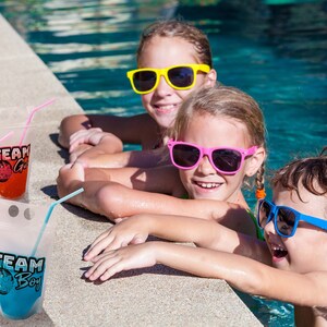 May include: Three children wearing sunglasses are sitting on the edge of a swimming pool. They are holding colourful pouches with straws, labelled "Team Girl" and "Team Boy".