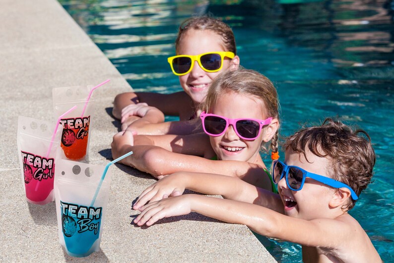 May include: Three children wearing sunglasses are sitting on the edge of a swimming pool. They are holding colourful pouches with straws, labelled "Team Girl" and "Team Boy".