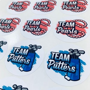 May include: Nine round white stickers with blue and pink designs. Six stickers feature the text "TEAM Pearls" with a pink pearl necklace design. Three stickers feature the text "TEAM Putters" with a blue golf bag and golf clubs design.