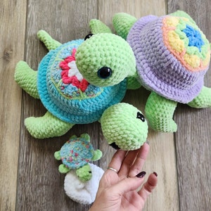 Puede incluir: Cuatro juguetes de tortuga de crochet en diferentes tamaños y colores. La tortuga más grande es morada con un caparazón arcoíris, la siguiente más grande es verde con un caparazón azul y rosa, la siguiente es verde con un caparazón blanco y la más pequeña es verde con un caparazón azul y rosa.