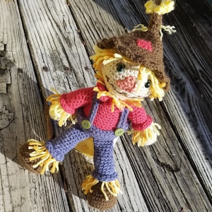 Op de afbeelding: Een gehaakte vogelverschrikkerpop met een bruine hoed, een rood shirt, een blauwe overall en geel strohaar. De pop zit op een houten oppervlak.