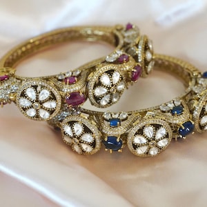 May include: Two ornate gold bangles with intricate detailing. Each bangle features a series of white, red, and blue gemstones set within a floral design, surrounded by smaller clear stones. The jewelry rests on a soft, light pink fabric.
