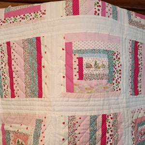 Könnte beinhalten: Eine weiße Steppdecke mit Patchwork-Muster in rosa, rot und blauen Blumenmustern. Die Steppdecke besteht aus Quadraten und Rechtecken, die Nähte sind sichtbar. Die Steppdecke hängt an einer Wand.