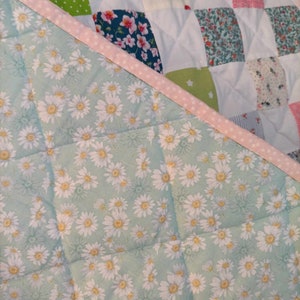 May include: A patchwork quilt with a green, white, and pink floral pattern. The quilt is made of squares of different fabrics and is quilted with a diamond pattern. The quilt is edged with a pink and white polka dot trim.